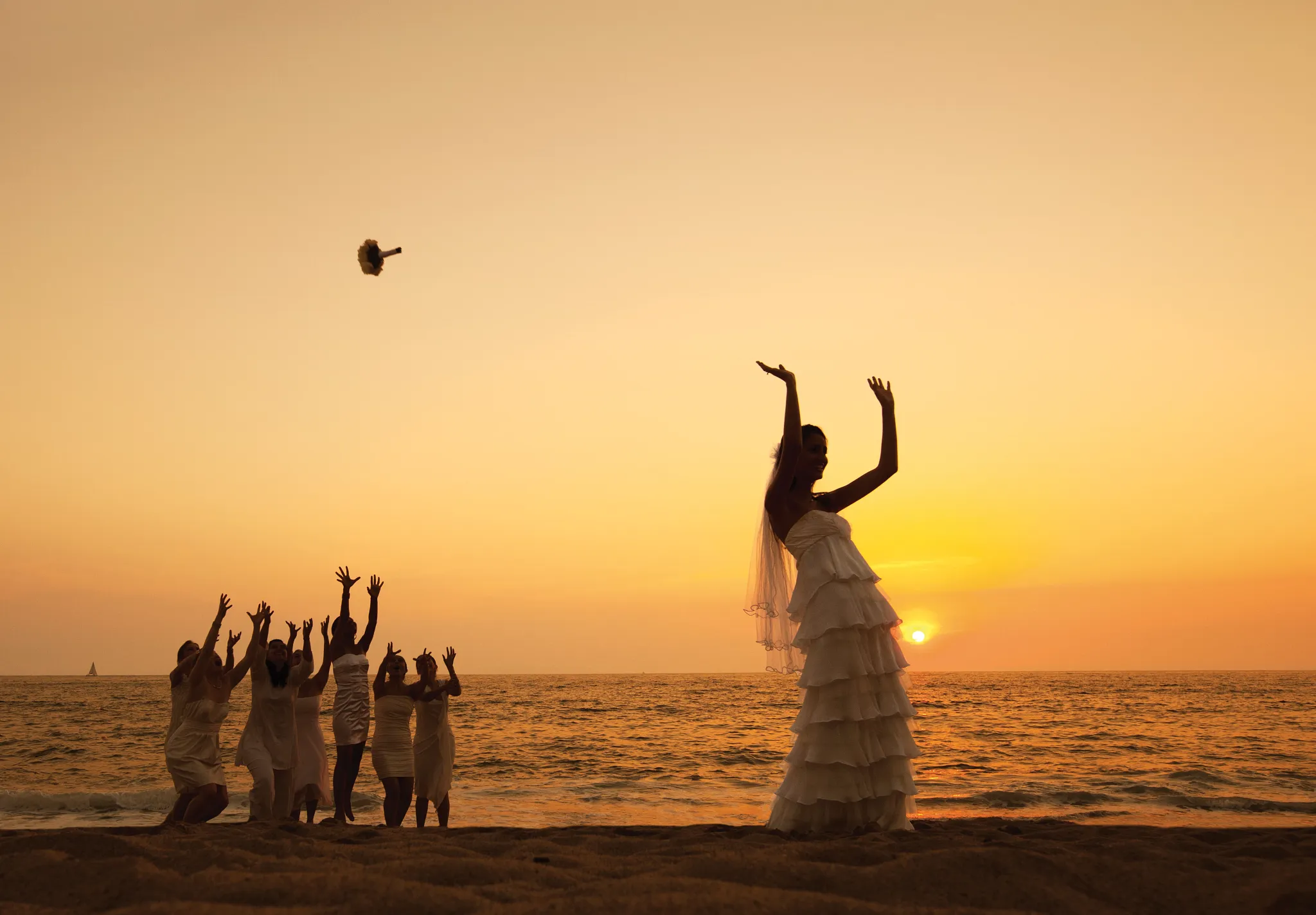 Bride tossing a bouquet to women on the beach at sunset